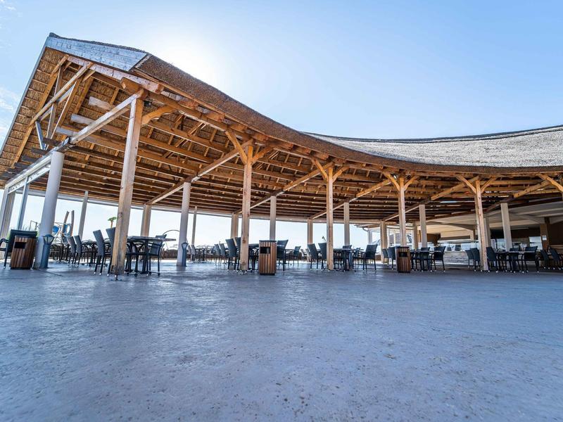 Estructura de madera abierta con sillas y mesas bajo un cielo claro junto a la piscina del hotel.