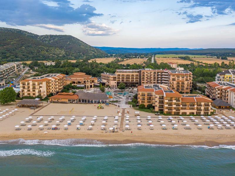 Vista de una playa con sombrillas y mar junto a un complejo con varios edificios y montañas al fondo.