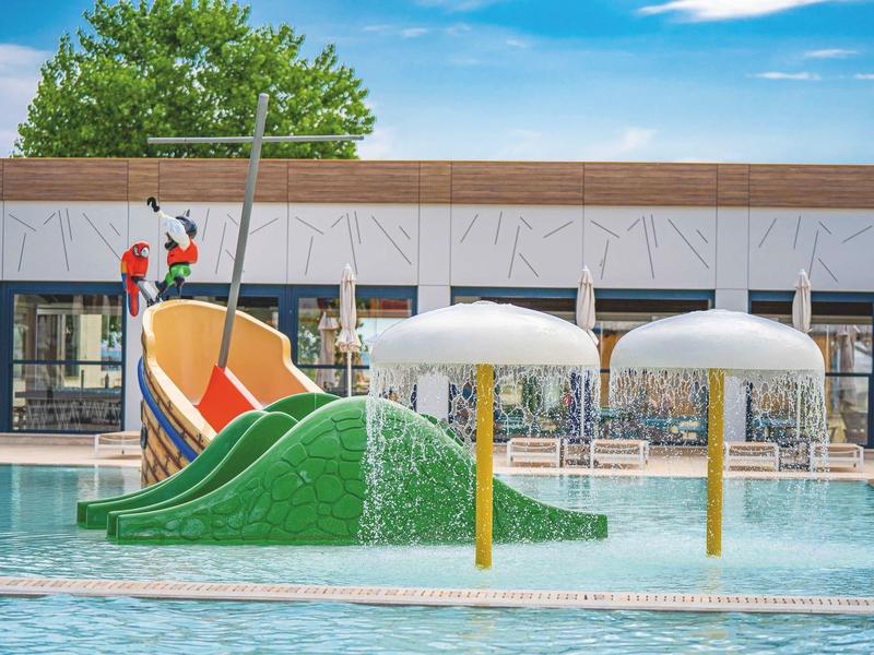 Parque acuático colorido con tobogán y fuentes en piscina bajo sol.