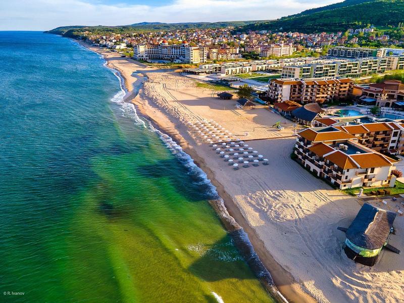 Vista aérea de un hotel en la amplia playa con mar verde y colinas al fondo.