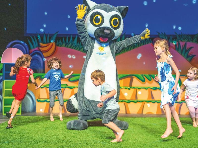 Niños juegan emocionados en el césped con una persona disfrazada de animal.