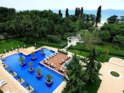 Großer blauer Pool mit Pflanzen und Liegen, umgeben von Bäumen und Blick auf das Meer.