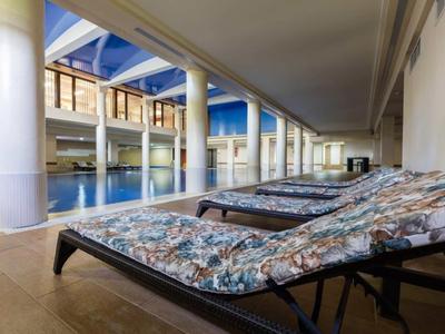 Innenbereich mit Schwimmbad, blauen Liegestühlen und großen weißen Säulen unter hellem Dach.