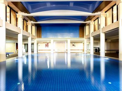 Helles Hallenbad mit blauem Wasser, hohen weißen Säulen und großem Fensterfronten an den Seiten.