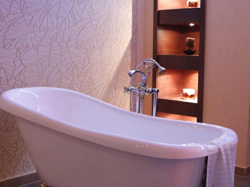 Freistehende weiße Badewanne mit Handtuch an der Seite vor beige gemusterter Wand und Regal mit Kerzen.