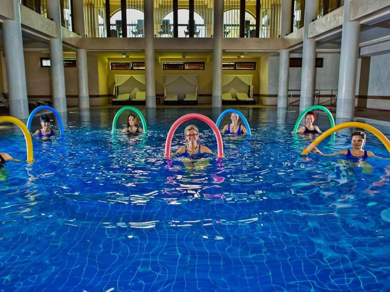 Mehrere Personen schwimmen mit bunten Schwimmnudeln im großen Hallenbad mit Säulen.