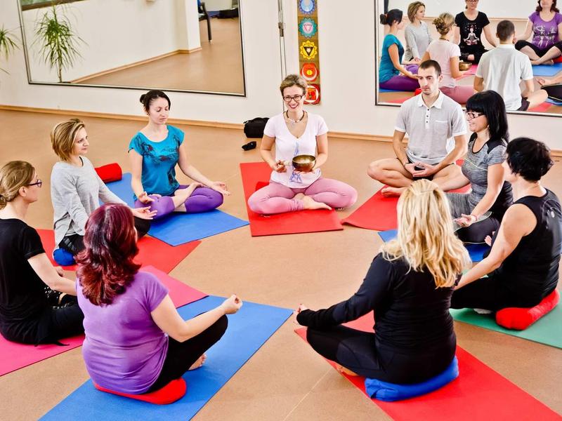 Sieben Personen sitzen im Kreis auf bunten Yogamatten und halten sich an den Händen.