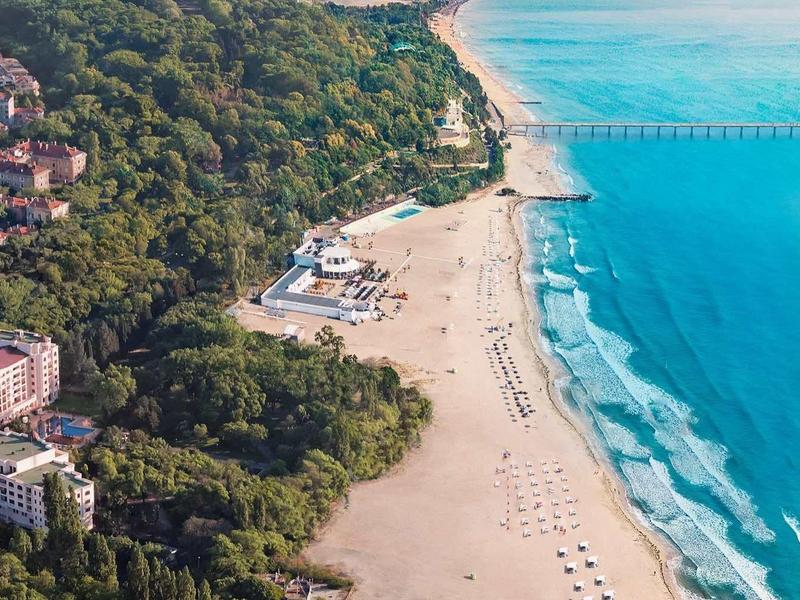 Langer Sandstrand mit blauem Meer, angrenzendem Wald und Stadtgebieten mit Gebäuden und Straßen.