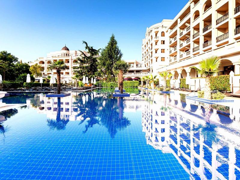 Großer Hotelpool mit blauer Kachel, Palmen und modernem Gebäude bei klarem Himmel.