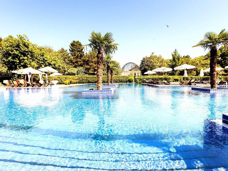 Großer blauer Pool mit Palmen, Sonnenschirmen und klarem Himmel im Hintergrund.