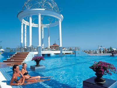 Dos mujeres descansan en la piscina de un resort con un pabellón blanco bajo un cielo azul claro.
