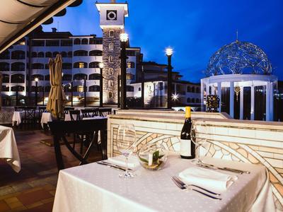 Terraza de hotel al atardecer con mesa puesta, botella de vino y pabellón iluminado al fondo.