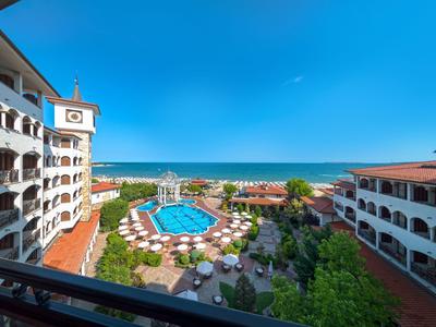Widok na hotel z basenem, parasolami i morzem w tle w słoneczny dzień.