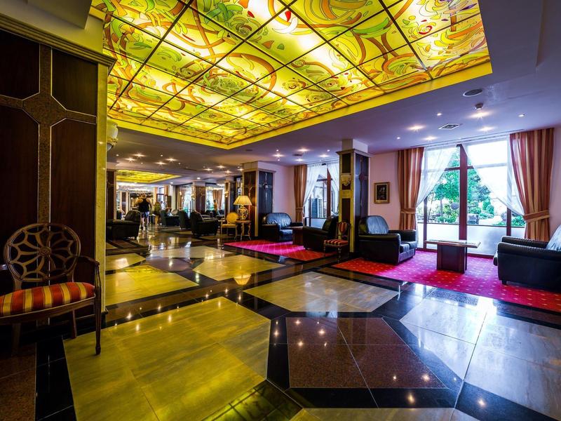 Elegante sala de hotel con techo de cristal, suelo de mármol y sofás negros con alfombras rojas.