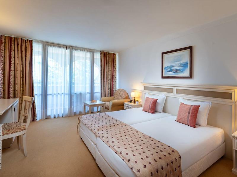 Habitación de hotel luminosa con cama doble, dos mesitas, escritorio, silla y gran ventana con cortinas.