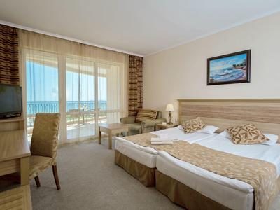 Bright hotel room with two single beds, desk, chair, and balcony overlooking the sea.