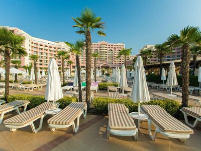 Hotel with pool and lounge chairs surrounded by palm trees under clear sky.