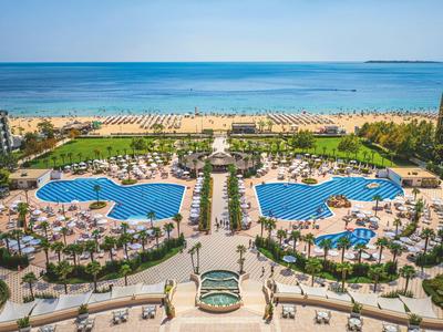 View of a hotel resort with two large pools, palm trees, and a beach in the background.