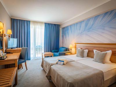Bright hotel room with two single beds, desk, and balcony with blue curtains.