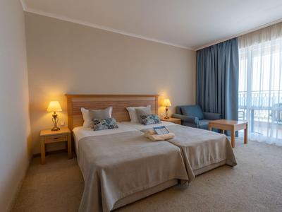 Hell eingerichtetes Hotelzimmer mit zwei Betten, Nachttischen, Stuhl und Balkon mit Vorhängen.