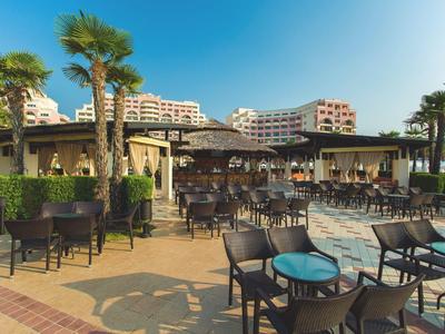 Hotel outdoor area with tables, chairs, palm trees, and apartment buildings in the background.