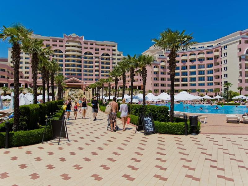 Large hotel with pool, palm trees, and people walking under a clear blue sky.