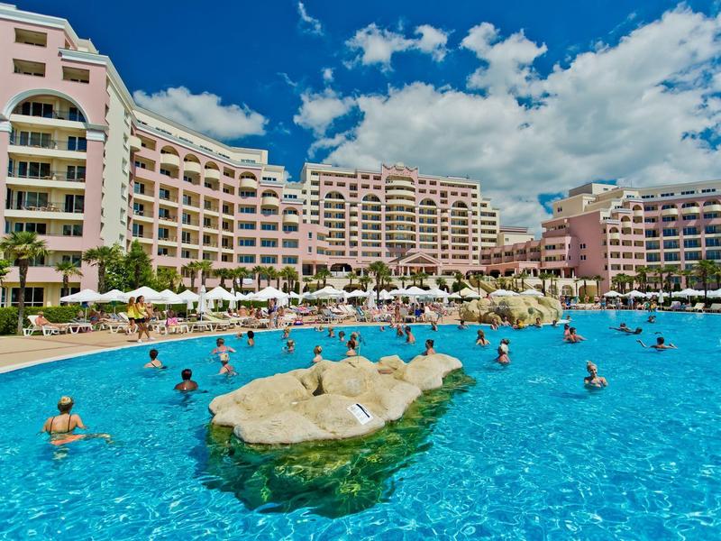 Large outdoor pool in front of a multi-story hotel under a blue sky with clouds.