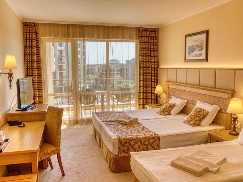 Bright hotel room with two single beds, balcony, and city view in daylight.
