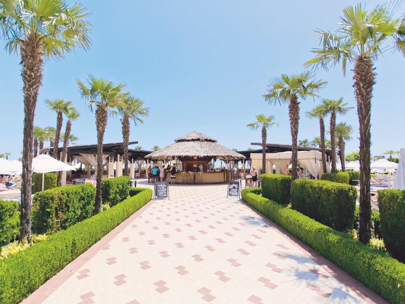 Wide pathway leading to a beach bar with palm trees and umbrellas under clear skies.
