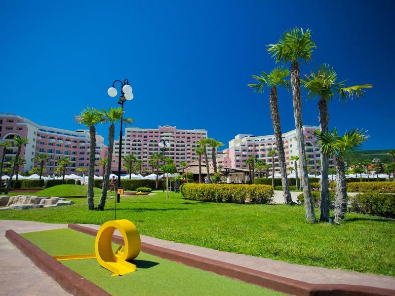 Green mini golf course with yellow obstacle and palm trees in front of large hotel buildings