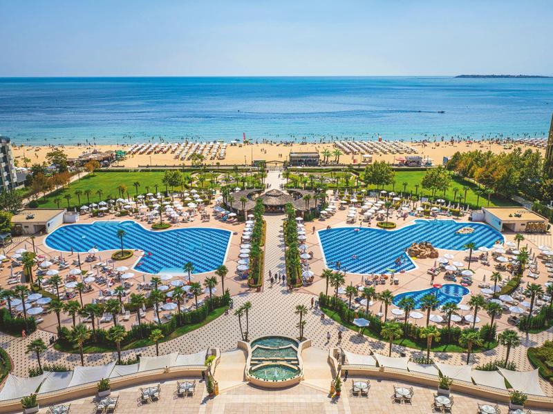 View of a hotel resort with two large pools, palm trees, and a beach in the background.