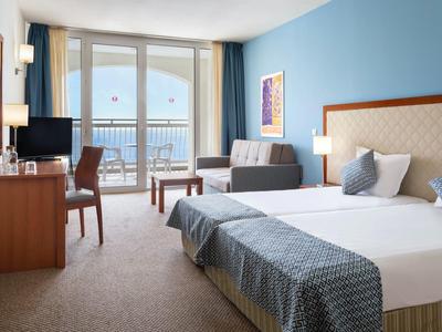 Bright hotel room with double bed, seating area, and balcony overlooking the sea.