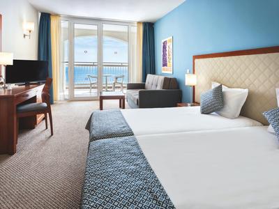 Bright hotel room with two beds, desk, sofa, and sea view through large windows.