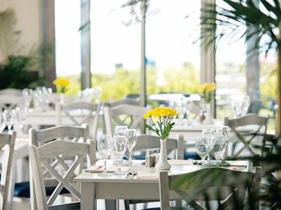 Brightly lit hotel dining area with white tables and chairs and yellow flowers in vases.