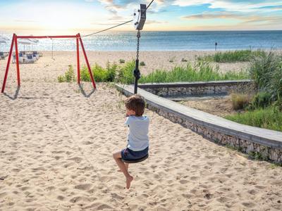 Child swings on a zip line on sandy beach overlooking the sea at sunset.