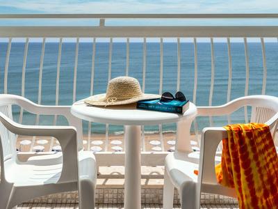 Balcony with white table and chairs, sea view, sun hat, book, and towel.