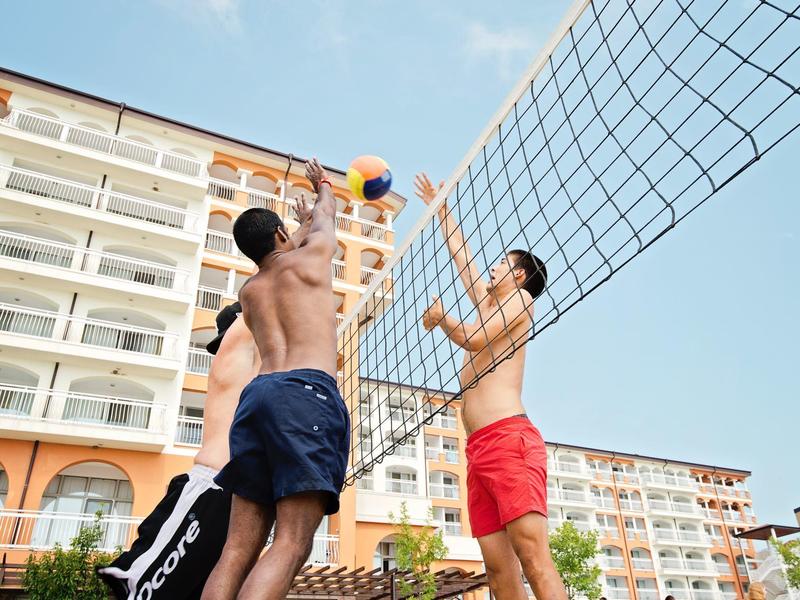 Two men playing beach volleyball in front of a hotel under clear sky.