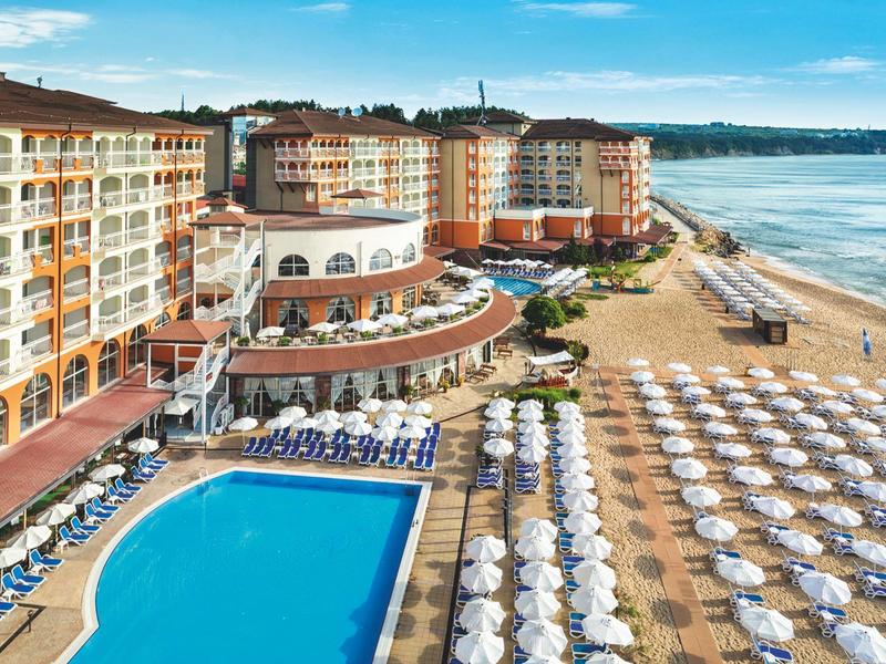 Large hotel with pool, lounge chairs, and umbrellas on the beach under a blue sky.