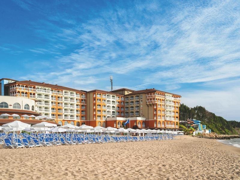 Large hotel building on sandy beach under blue sky with rows of sun loungers.