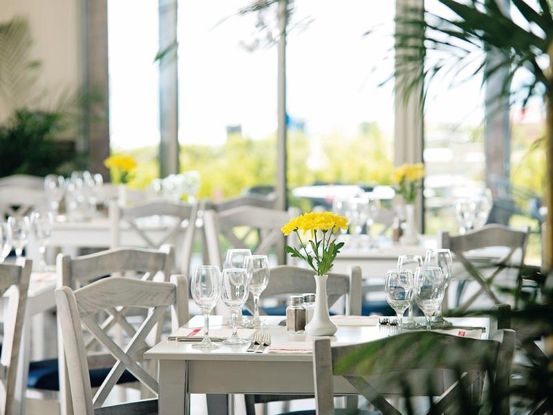Brightly lit hotel dining area with white tables and chairs and yellow flowers in vases.