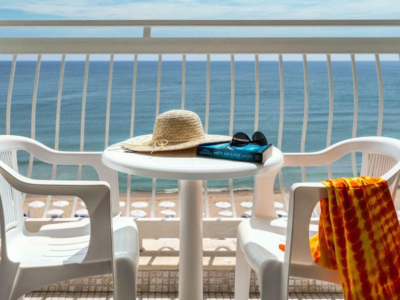 Balcony with white table and chairs, sea view, sun hat, book, and towel.