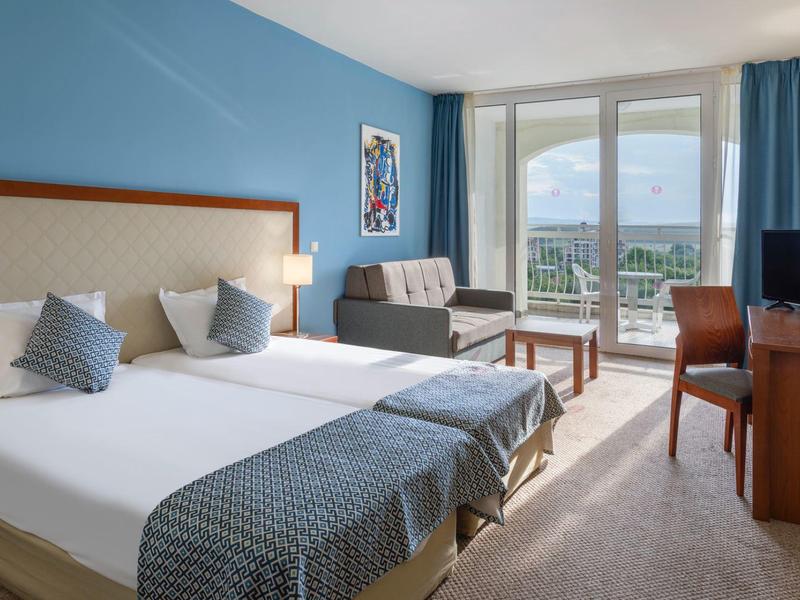 Modern hotel room with two single beds, sofa, and balcony overlooking green landscape.