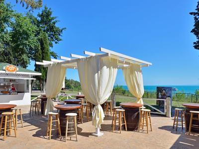 Strandcafé mit weißen Vorhängen, Holzstühlen und Tischen, blauer Himmel und grüner Baumkulisse.
