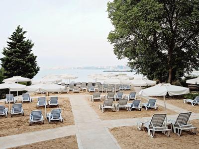 Strand mit vielen Liegestühlen, weißen Sonnenschirmen, Bäumen und ruhigem Meer im Hintergrund.
