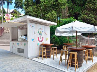 Kleines weißes Kiosk mit Holztheke, hohen Stühlen und weißem Sonnenschirm im Freien vor Bäumen.