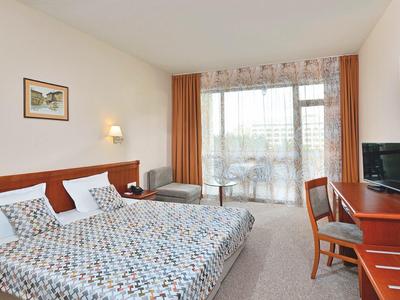Helles Hotelzimmer mit Doppelbett, Schreibtisch, TV, Balkon und orangefarbenen Vorhängen.