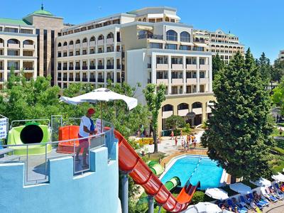 Hotel mit Wasserrutsche, Pool und Sonnenschirmen, umgeben von Bäumen und blauem Himmel.