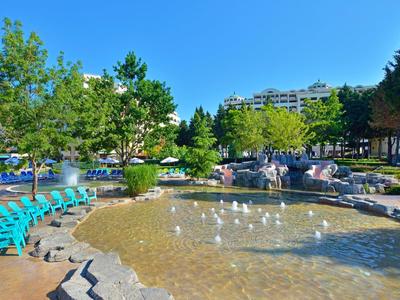 Stadtpark mit klarem Teich, Enten, grünen Bäumen, Sitzbänken und modernen Gebäuden im Hintergrund.