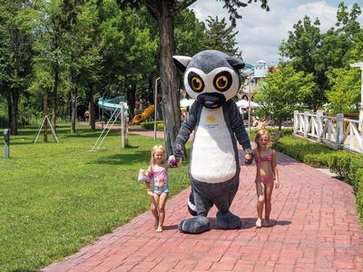 Zwei Kinder laufen auf einem roten Gehweg neben einer Person im Pinguinkostüm in einem Park.