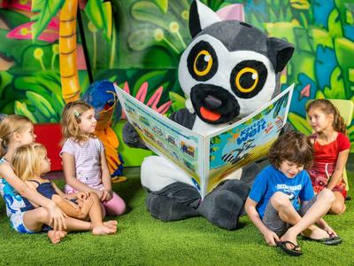 Fünf Kinder sitzen im Kreis auf grünem Rasen, hören einem großen Lemuren-Maskottchen zu, das ein Bilderbuch hält.